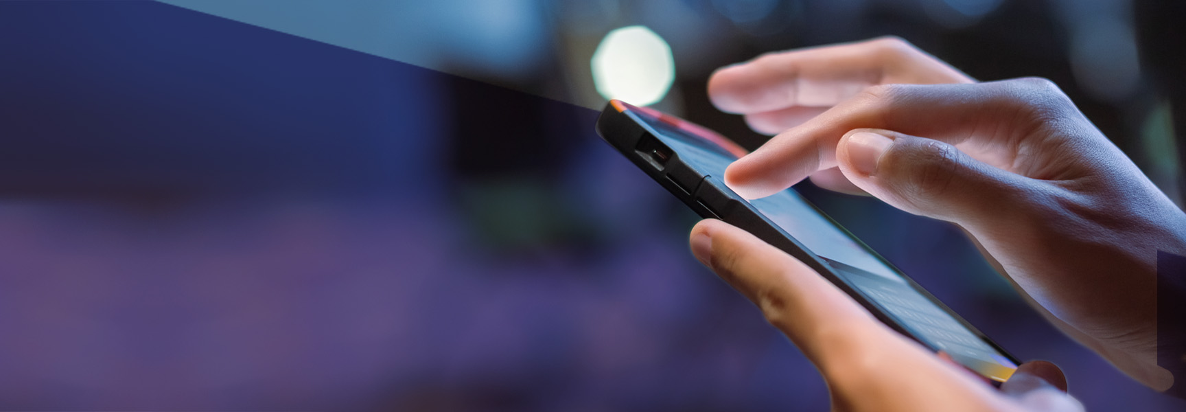 Close-up of hands holding a smartphone with fingers tapping on the screen. The background is blurred with dark blue and purple hues, suggesting evening or night time.