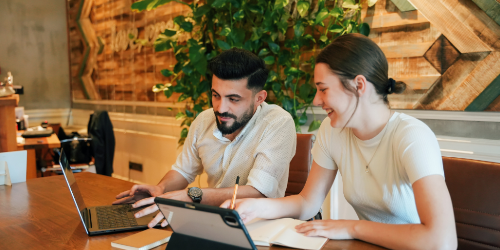 Two colleagues sit at a wooden table, collaborating. The man uses a laptop, while the woman takes notes with a pen and notebook. They appear focused and engaged, with a plant and wooden decor in the background.