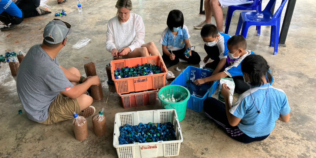 A group of people, including adults and children, sit on the ground organizing colorful plastic caps into bins. Several people wear blue uniforms, and others wear casual clothing. Some children are wearing face masks.