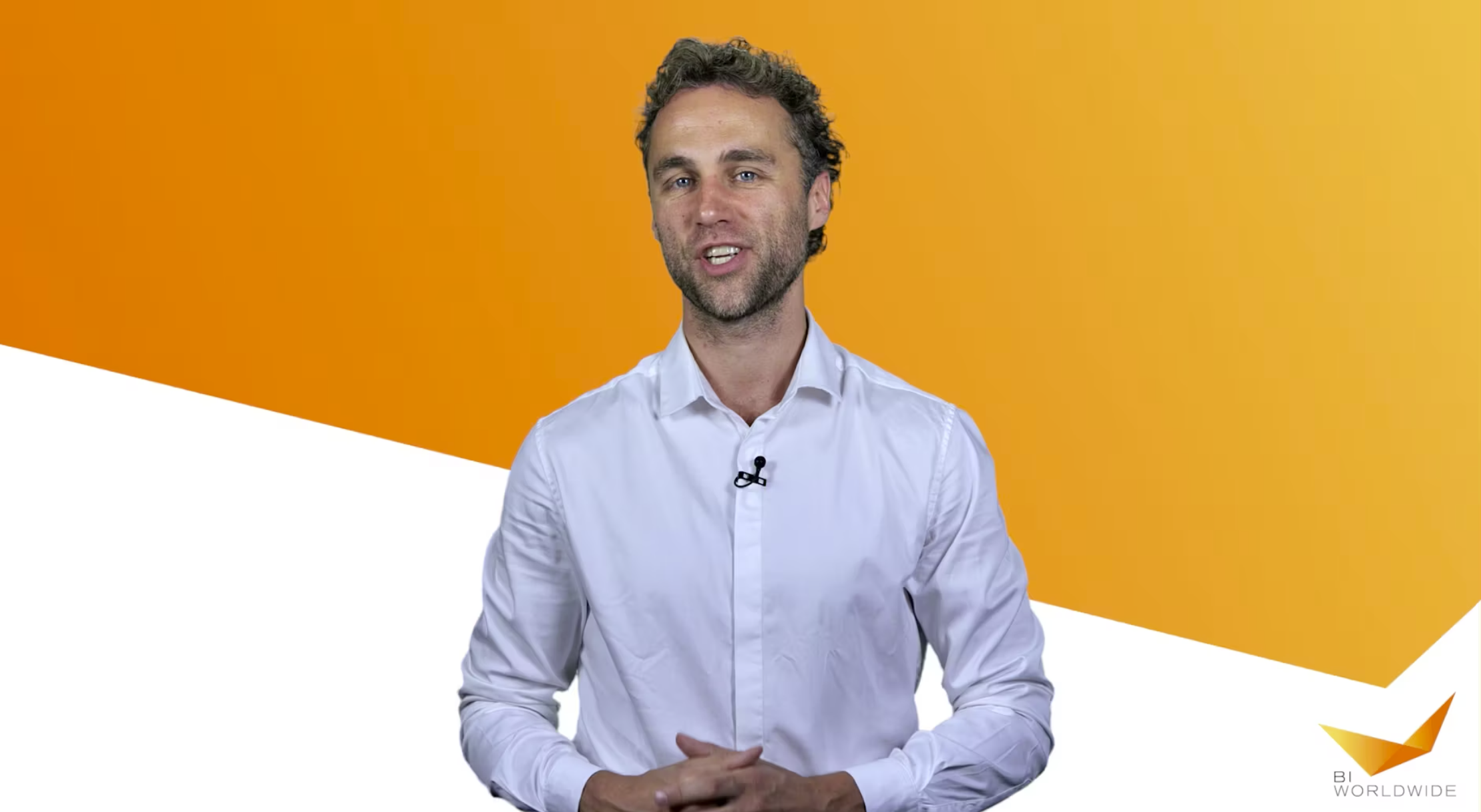 A man with curly hair is wearing a white shirt and smiling. He stands against an orange and white geometric background. A logo in the bottom right corner reads BI WORLDWIDE.