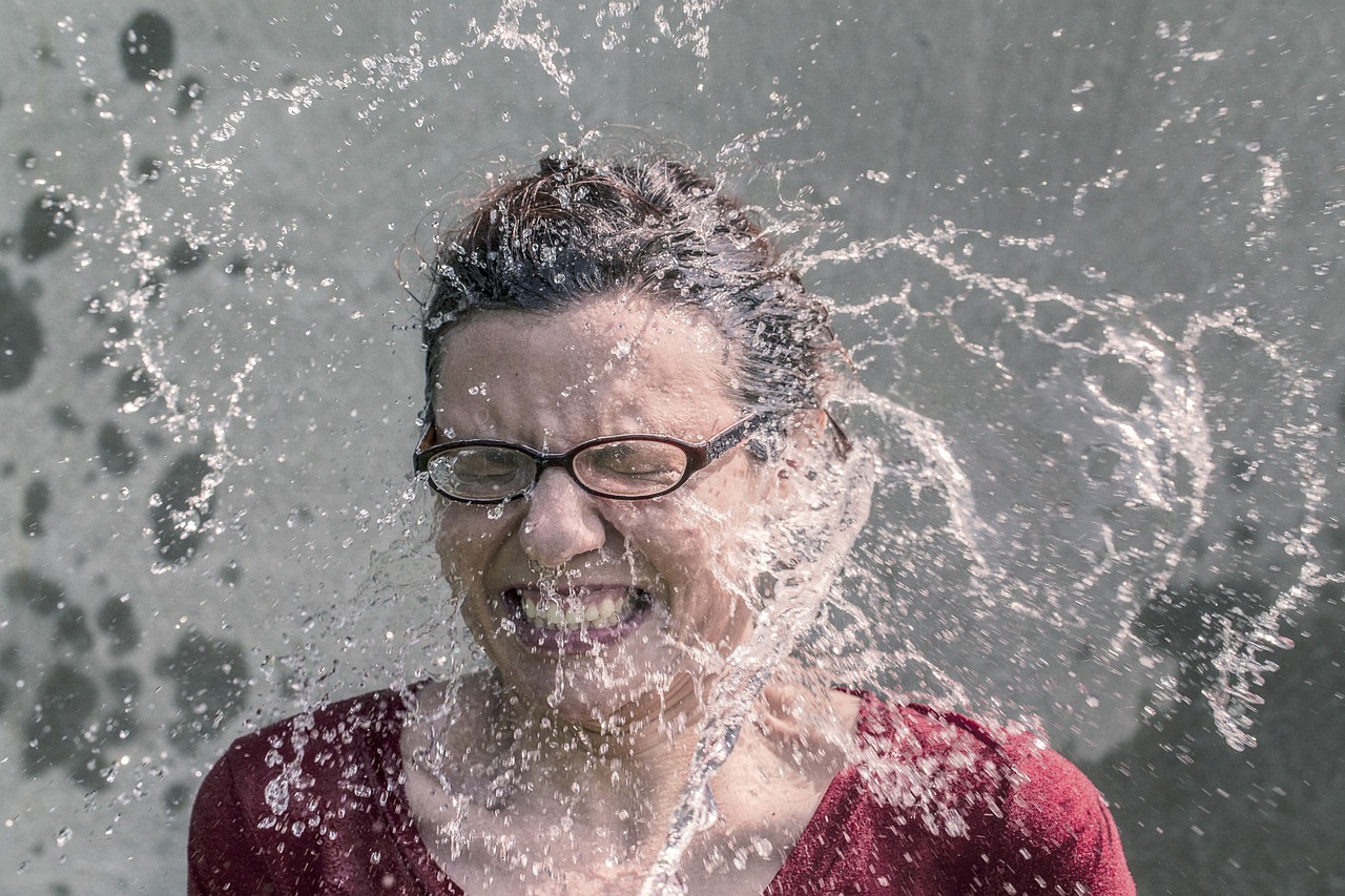 A person with glasses and a red top is caught mid-expression as water splashes dramatically onto their face against a gray background.