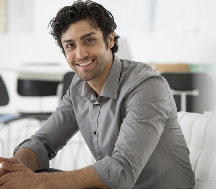 Smiling man with dark curly hair and a beard, wearing a gray button-up shirt, sits on a white sofa in a bright, modern room with blurred background elements, including black chairs and tables.
