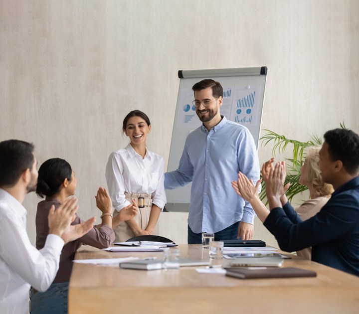 A man and woman stand smiling next to a presentation easel with charts. Four colleagues at a table applaud them. The room has a plant and a bright, professional atmosphere.