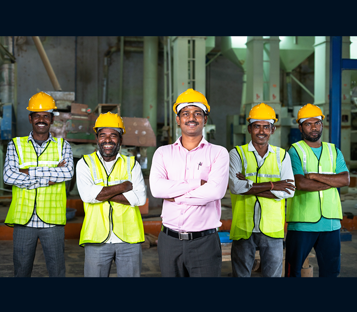 Five men wearing yellow hard hats and reflective vests stand with arms crossed in an industrial setting. The central man wears a pink shirt without a vest, smiling confidently. Machinery and equipment are visible in the background.
