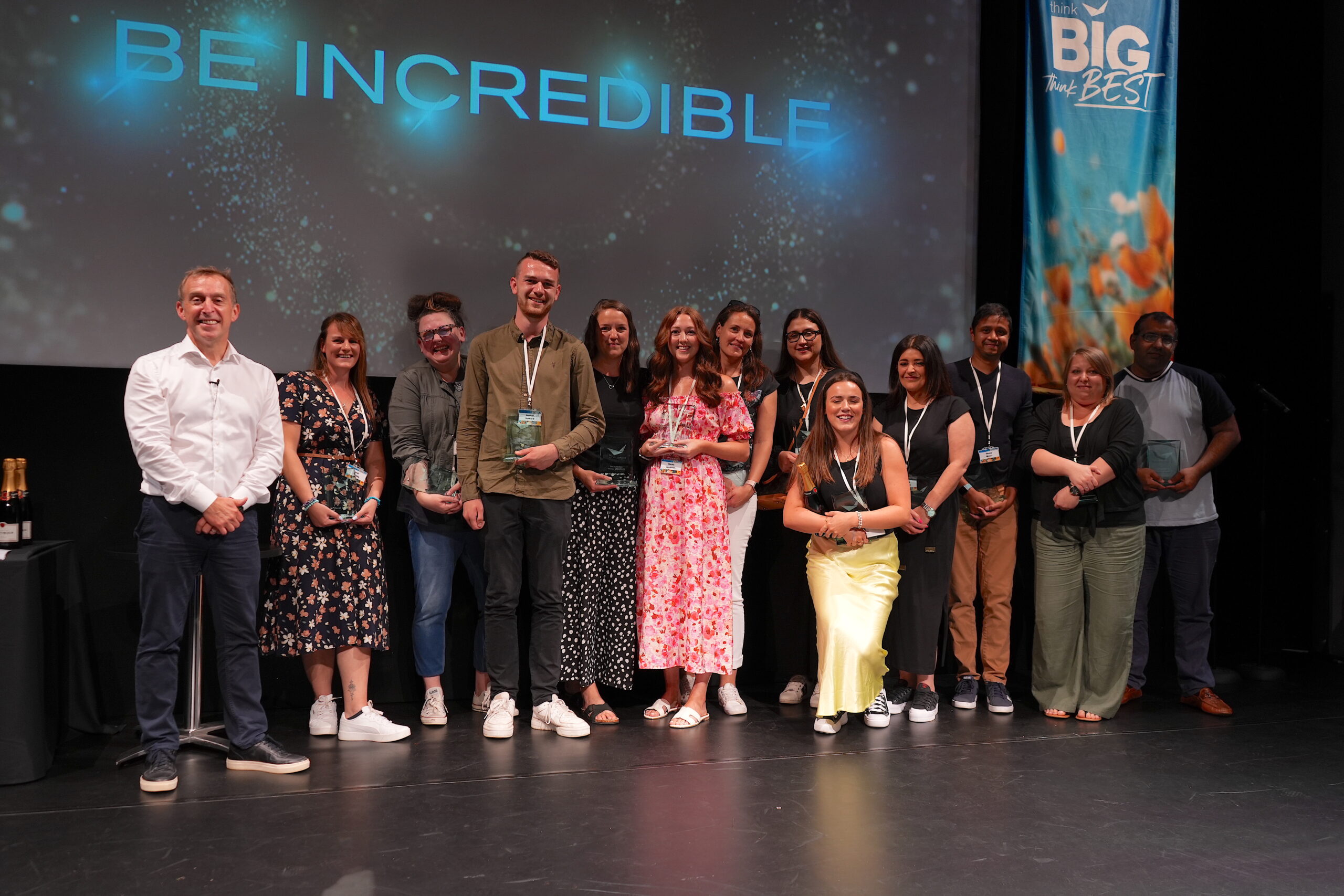 A group of people stand on stage smiling, holding awards. Behind them is a screen displaying the phrase Be Incredible. They are at an event with a sign that reads BIG the BEST.