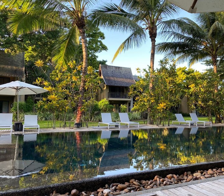 A luxury Mauritius oasis awaits, with a serene outdoor pool surrounded by palm trees and lounge chairs under umbrellas. In the background, tropical plants and quaint wooden-roofed cabins set the stage for an exclusive event for a global analytics company. Pebble-bordered pool and wooden deck complete this idyllic escape.