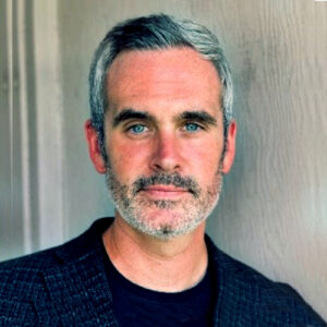 A man with short gray hair and a beard stands in front of a textured wall. He is wearing a dark blazer over a black shirt, looking directly at the camera with a neutral expression.