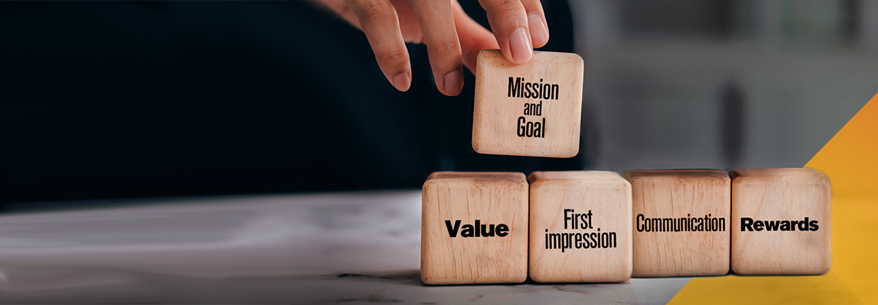 A hand places a wooden block labeled Mission and Goal on top of a row of blocks with the words Value, First Impression, Communication, and Rewards. The background is blurred with a yellow triangle on the right edge.