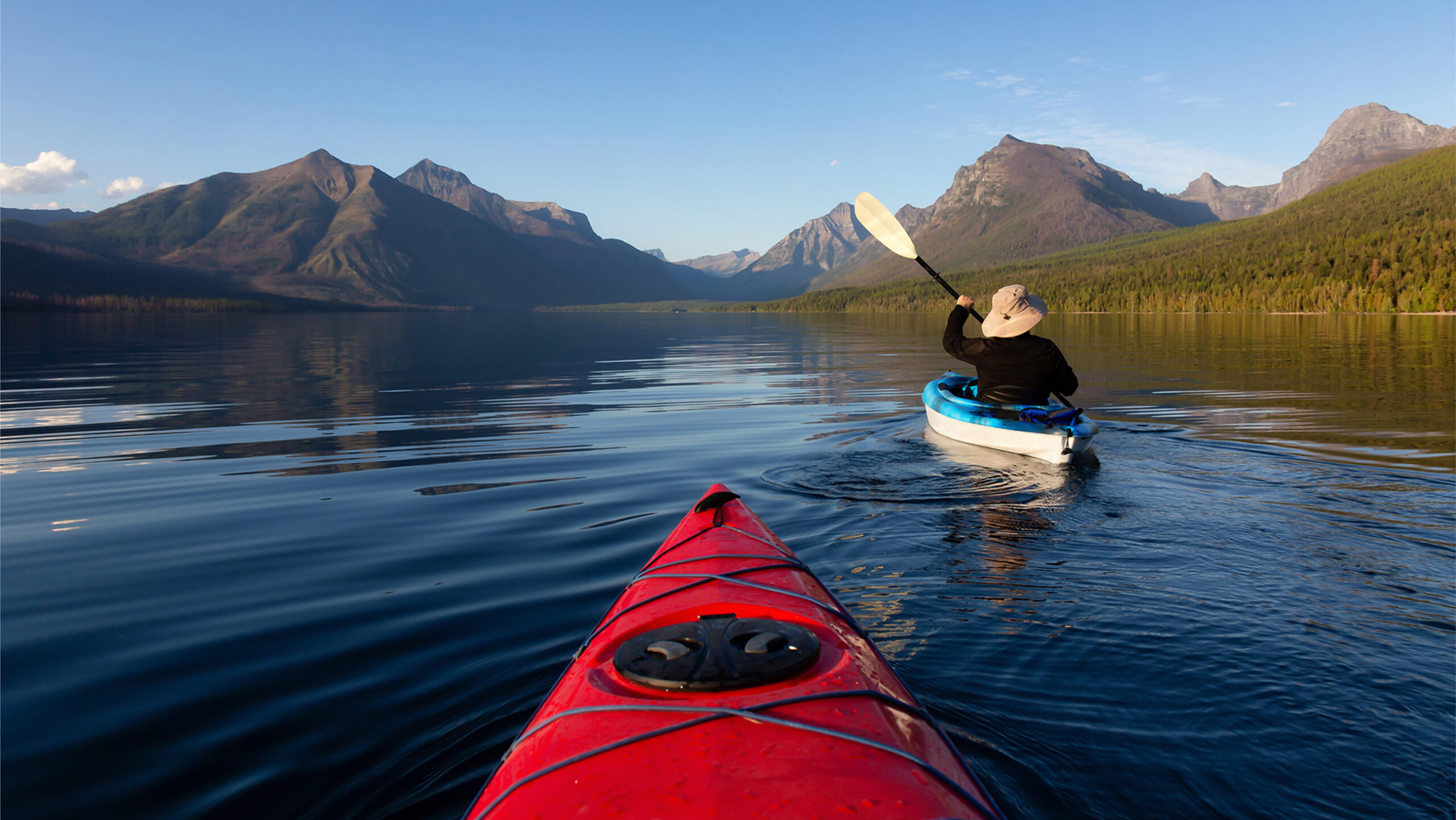 A person paddles a blue kayak on a calm lake, surrounded by mountains under a clear blue sky. The tip of a red kayak is visible in the foreground, enhancing the perspective of the serene natural setting.