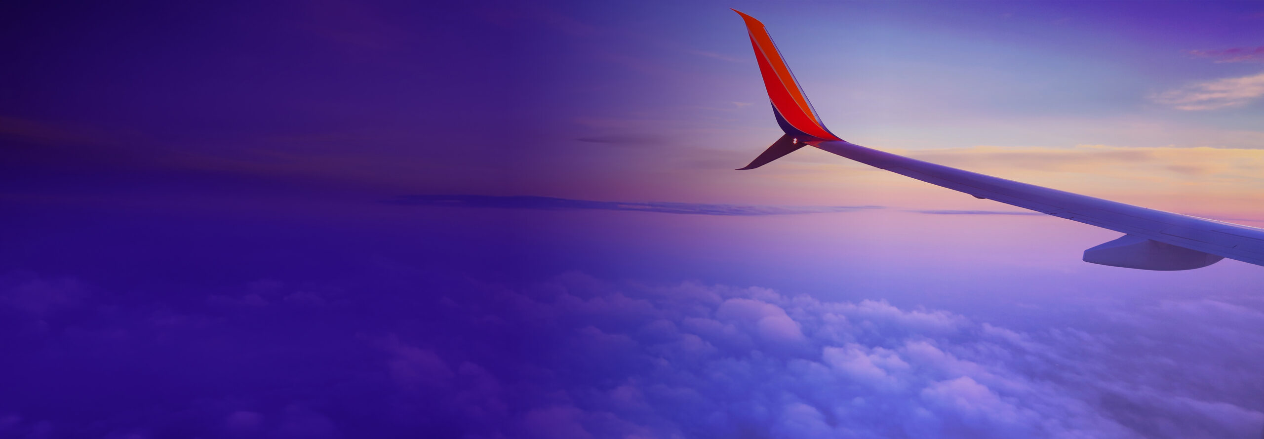 View from an airplane window showing a wing with a colorful sky in the background. The sky transitions from deep purple to warm orange and pink hues, reflecting a serene sunset or sunrise above fluffy clouds.