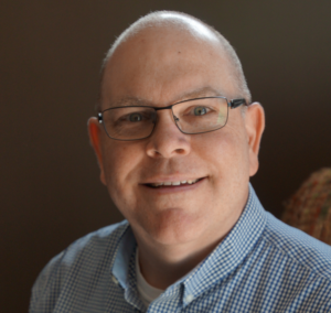 Smiling man with glasses and a short haircut, wearing a blue checkered shirt, sitting in a softly lit room.