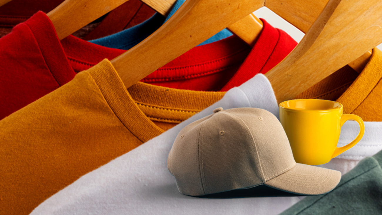 Close-up of colorful t-shirts on wooden hangers, featuring red, blue, yellow, and white shirts. In the foreground, a beige baseball cap and a bright yellow mug are placed together.