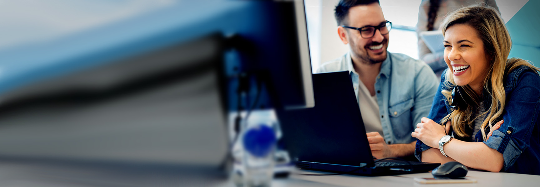 Two people smiling and talking while looking at a computer screen. The man is wearing glasses and a light blue shirt. The woman is wearing a denim jacket and headphones. They are in a modern office setting with blurred background elements.