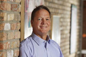 A man in a light blue shirt stands against a brick wall. He is smiling and looking at the camera. The background includes framed pictures on the wall.