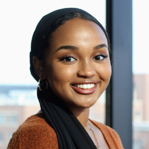 A woman wearing a black headscarf and orange sweater smiles at the camera. She has hoop earrings and a necklace. The background shows a window with an urban cityscape.