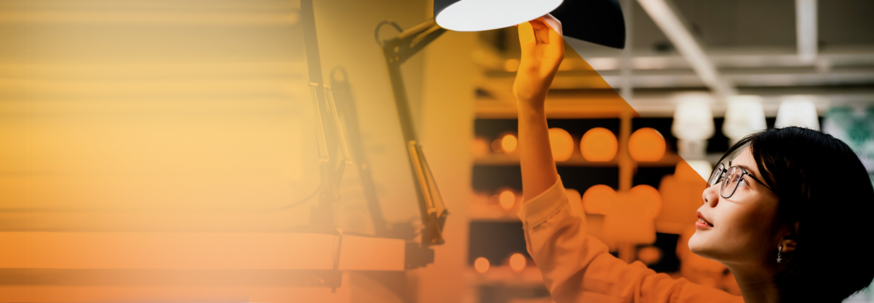 A person wearing glasses is reaching up to adjust a desk lamp, surrounded by a warmly lit interior. The right side of the image is in focus, while the left side features a soft orange gradient.