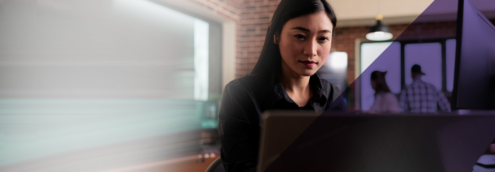 A woman sits at a desk working on a laptop in an office setting. The background is blurred, showing two people walking and details of the office space. She is focused on her work, with soft lighting illuminating her face.