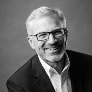 Smiling man with short gray hair, beard, and glasses, wearing a dark suit and white shirt, against a plain background. Black and white photo.