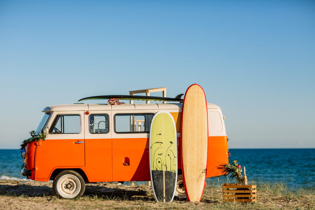 A vintage orange van is parked on a beach, with two surfboards resting against it. The ocean is visible in the background under a clear blue sky. A crate with flowers is placed next to the van, adding a decorative touch.