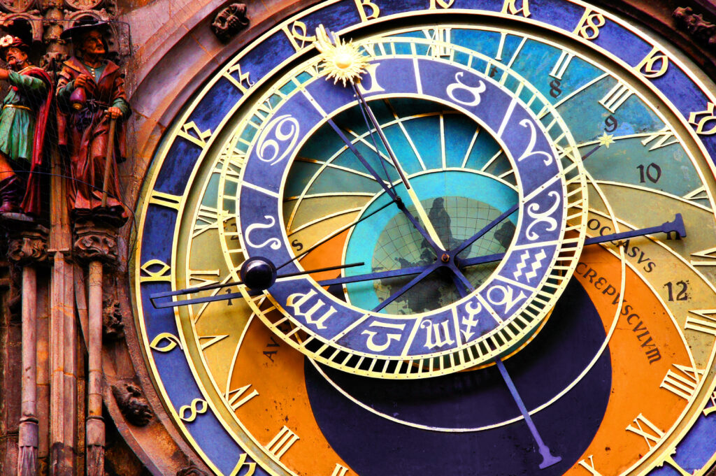 Close-up of an ornate astronomical clock face featuring zodiac signs and Roman numerals with vibrant blue and gold detailing. Sculpted figures are visible on the left side, adding an artistic touch to the intricate design.