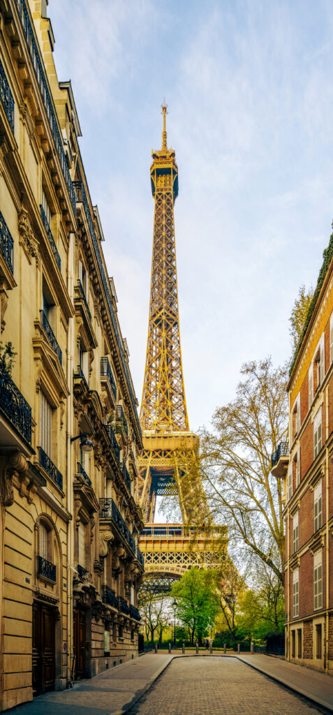 The Eiffel Tower stands tall in the background, framed by two rows of elegant old buildings. Leafy trees line the quiet street leading towards the tower under a clear blue sky.