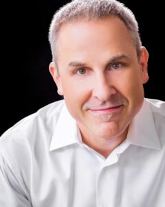 Portrait of a man with short gray hair, wearing a white shirt, and smiling. He is positioned against a solid black background, looking directly at the camera.