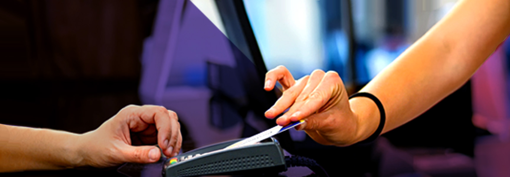 Two hands are visible at a counter. One hand is holding a credit card and inserting it into a card reader. The other hand is resting nearby. A monitor is partially visible in the background.