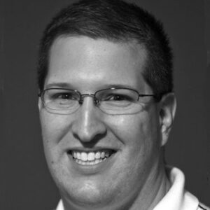 A black-and-white portrait of a person wearing glasses and smiling, with short hair and a light-colored, collared shirt, set against a plain, dark background.