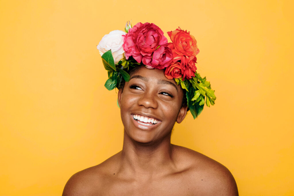 A person smiling brightly wears a vibrant flower crown with red and white roses, set against a solid yellow background.