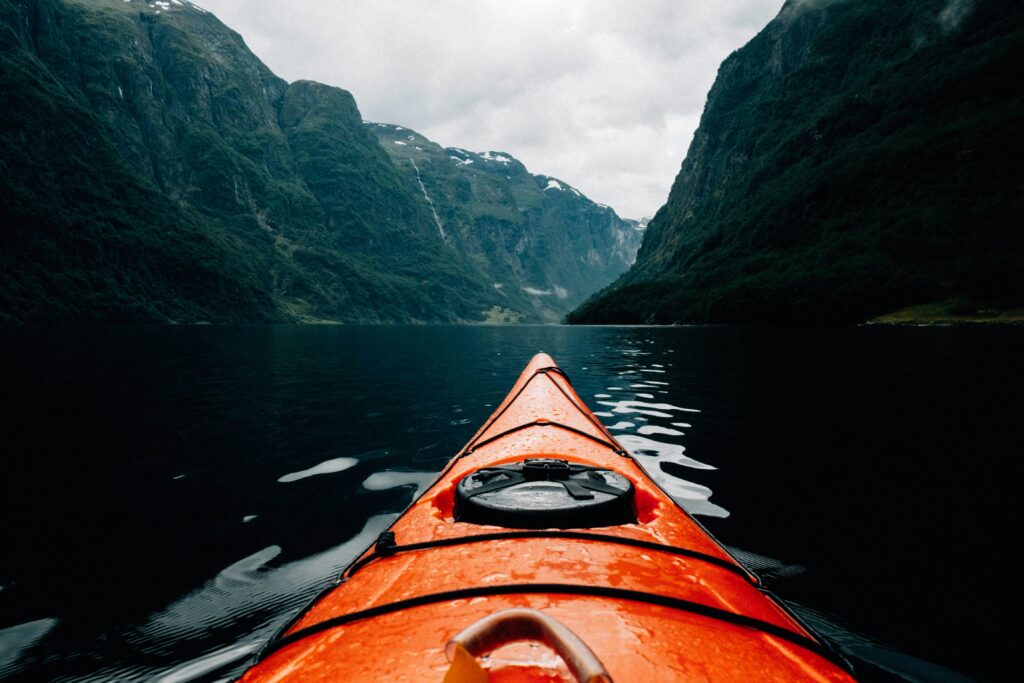 View from the seat of an orange kayak on a calm, dark lake surrounded by steep, green mountain cliffs under a cloudy sky.