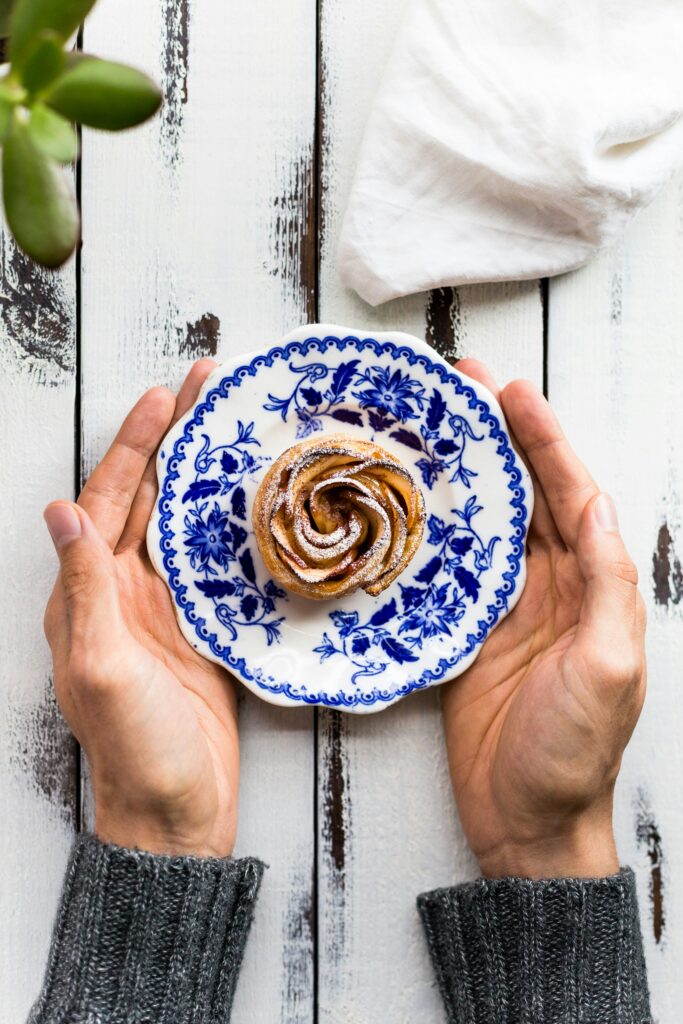 Hands holding a blue and white floral-patterned plate with a cinnamon roll dusted with powdered sugar on a white wooden table. A green plant and white cloth are partially visible in the background.