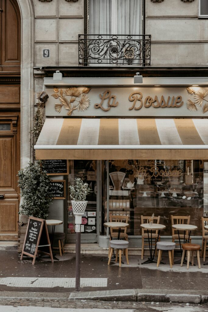 A charming Parisian café with a striped awning labeled La Bossue. Wooden chairs and tables are visible inside through large windows. A chalkboard menu stands outside, next to a small potted plant, creating a cozy street-side ambiance.