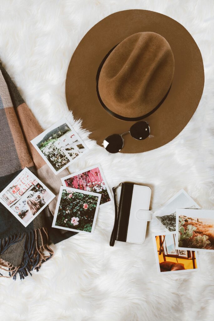 A brown hat and round sunglasses rest on a white fluffy surface. Nearby are several instant photos, a folded plaid scarf, and a white wallet with a black strap. The photos depict various scenes, including flowers and landscapes.