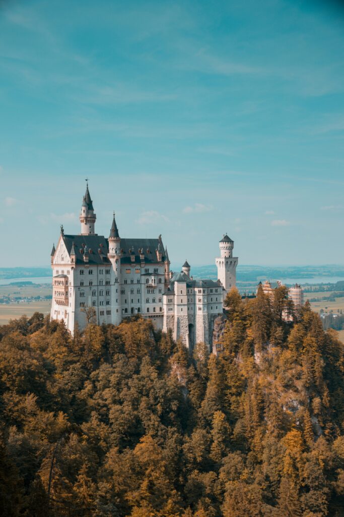 Scenic view of a large, ornate castle with tall spires and turrets, perched atop a forested hill under a clear blue sky. The landscape stretches out in the background with fields and distant mountains.