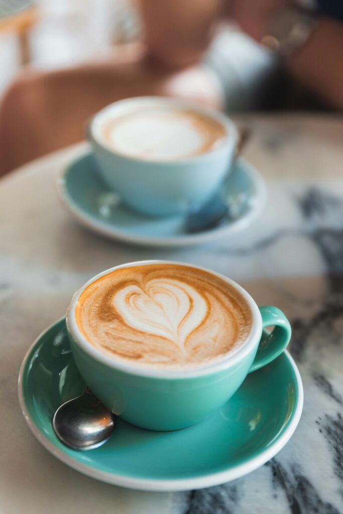 Two cups of coffee with heart-shaped latte art on teal saucers are placed on a marble table. A spoon rests on the saucer of the closer cup. Soft focus background includes another person and part of a table.