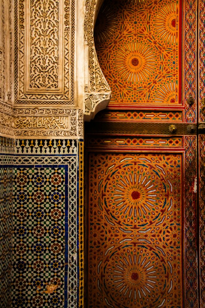 Intricate wall art featuring vibrant geometric patterns in red, orange, and blue hues. The design includes detailed mosaic tile work and ornate carved stone, typical of traditional Islamic architecture.