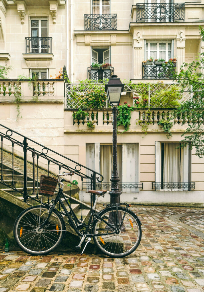 A bicycle is parked against a vintage lamppost beside a staircase leading to an ornate building with balconies. The cobblestone courtyard is surrounded by lush greenery and window boxes with flowers. The scene is serene and picturesque.