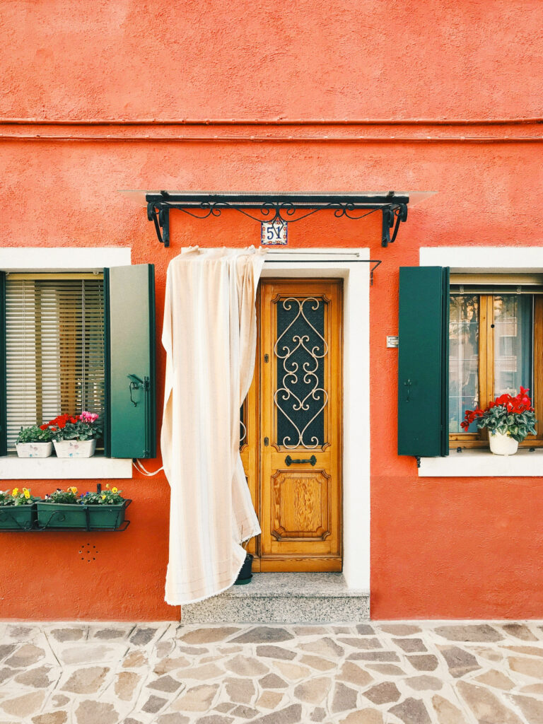 A vibrant orange house with green shutters and a wooden door. A light curtain partially covers the door, which has an ornate iron design. Flower boxes with colorful blooms adorn the windowsills on each side. The house number 59 is displayed above.