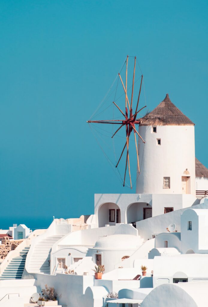 A picturesque scene of a white windmill with a thatched roof on a hillside in Santorini, Greece. The building is surrounded by whitewashed structures with blue accents, under a clear blue sky, evoking a serene Mediterranean atmosphere.