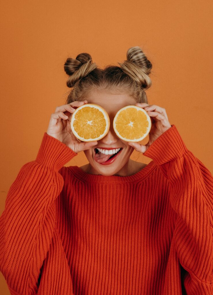 A person with two buns in their hair holds orange slices over their eyes, sticking out their tongue. They are wearing a bright orange sweater against a matching orange background, creating a playful and vibrant scene.