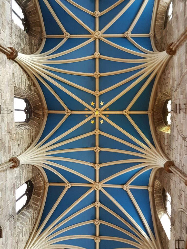 The image shows a vaulted ceiling of a cathedral with a pattern of white ribs forming star-like shapes on a blue background. Small golden stars are at the center. The surrounding walls are made of stone with arched windows.