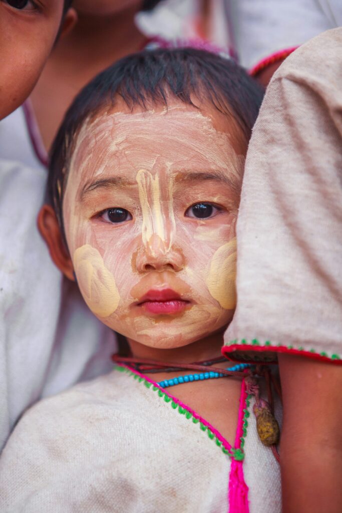 A young child with traditional thanaka paste design on their face gazes at the camera. The child is wearing a white garment with colorful trim. They are surrounded closely by other people, creating a warm, intimate atmosphere.